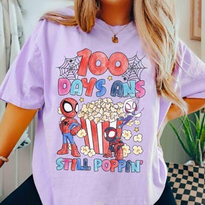 May include: A light purple t-shirt featuring a colorful graphic celebrating 100 days of school. The design includes the text "100 Days And Still Poppin" with Spider-Man characters, popcorn, and spiderwebs. A fun and festive design for students.