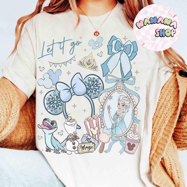 Może przedstawiać: Kremowa koszulka z fantazyjnym wzorem z napisem "Let it go". Projekt zawiera ilustracje Elsy, Olafa i innych element&oacute;w tematycznych z Krainy Lodu w odcieniach niebieskiego i białego. Występuje r&oacute;wnież opaska na głowę w kształcie kokardy i korona.