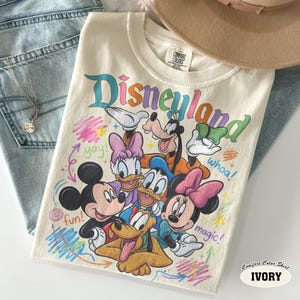 K&ouml;nnte beinhalten: Ein elfenbeinfarbenes T-Shirt mit einem bunten Grafikdruck von Mickey Mouse, Minnie Mouse, Donald Duck, Daisy Duck, Goofy und Pluto. Der Text Disneyland ist oben aufgedruckt, erg&auml;nzt durch die W&ouml;rter yay, whoa, fun und magic.