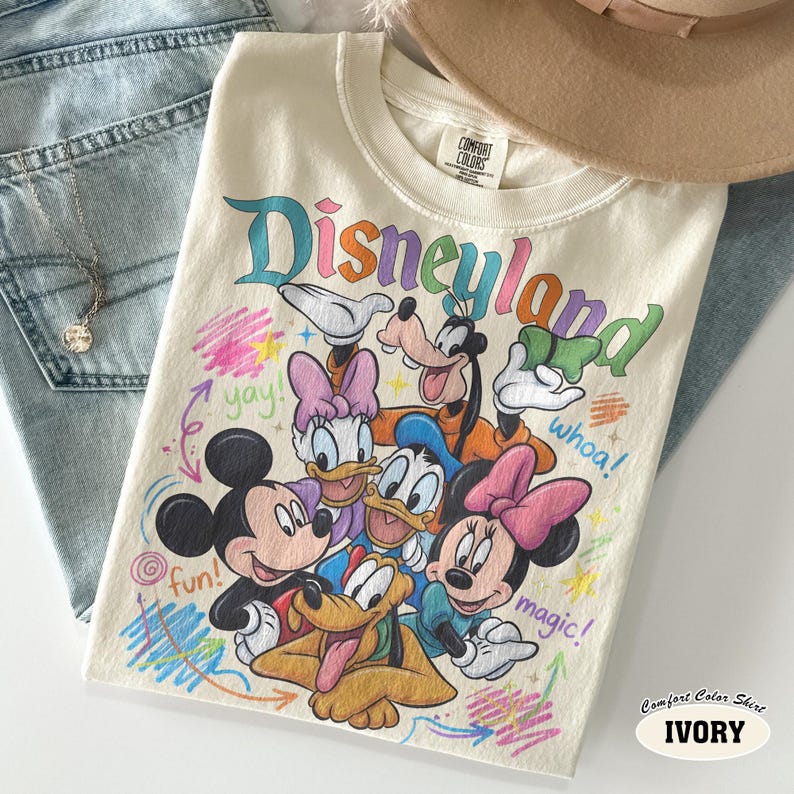 K&ouml;nnte beinhalten: Ein elfenbeinfarbenes T-Shirt mit einem bunten Grafikdruck von Mickey Mouse, Minnie Mouse, Donald Duck, Daisy Duck, Goofy und Pluto. Der Text Disneyland ist oben aufgedruckt, erg&auml;nzt durch die W&ouml;rter yay, whoa, fun und magic.