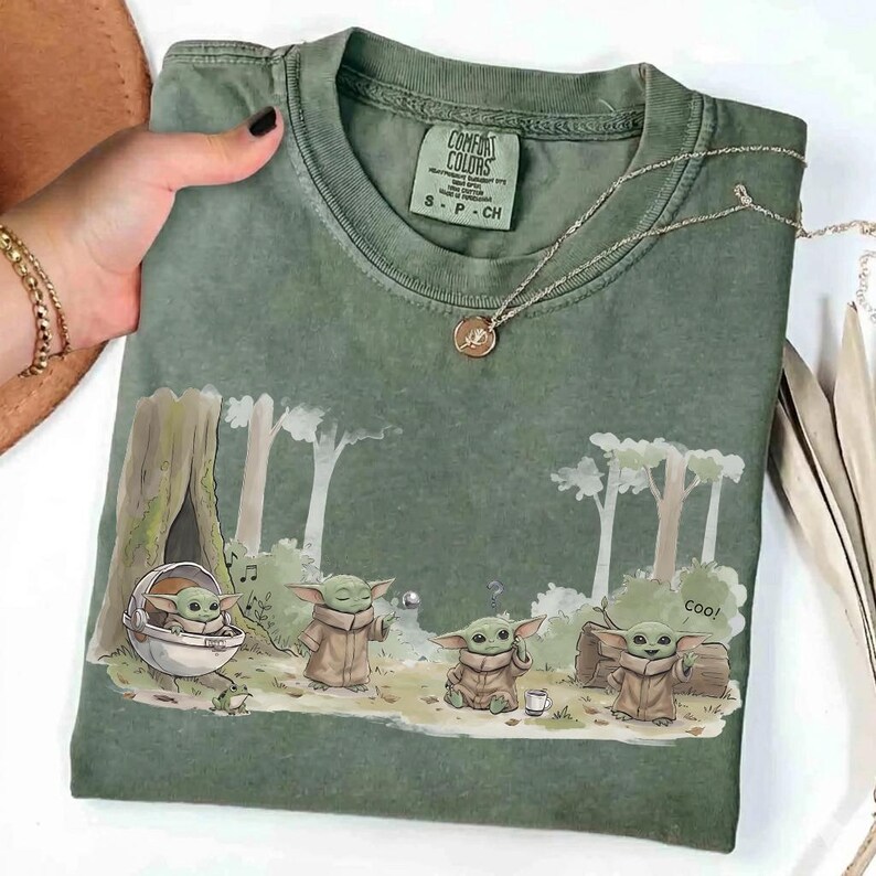 May include: Green Comfort Colors t-shirt featuring a cartoon illustration of a character from a popular science fiction series. The graphic shows the character in various poses in a forest setting. A silver necklace with a pendant is also visible.