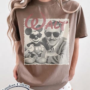 Pu&ograve; includere: T-shirt color espresso con un'immagine in bianco e nero di Walt Disney e Topolino con occhiali da sole. La parola "Walt" &egrave; stampata in rosso sopra l'immagine. La maglietta &egrave; di Comfort Colors.