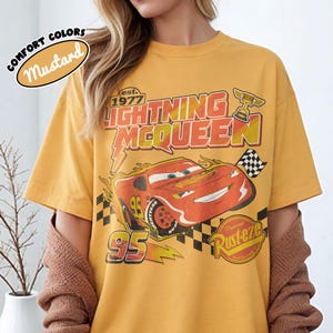 May include: Mustard-coloured t-shirt with a Lightning McQueen graphic from the film Cars. The design includes the text "Lightning McQueen," "95," and "Rusteze." The shirt has short sleeves and a crew neck.