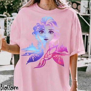 May include: Pink t-shirt featuring a colorful graphic of a woman's face with a landscape design. The graphic includes shades of blue, purple, and pink. The word "Blossom" is printed in black at the bottom left of the shirt.