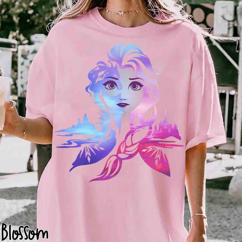 May include: Pink t-shirt featuring a colorful graphic of a woman's face with a landscape design. The graphic includes shades of blue, purple, and pink. The word "Blossom" is printed in black at the bottom left of the shirt.