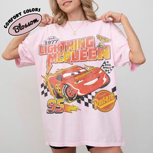 May include: A pale pink t-shirt with a graphic of Lightning McQueen from the Cars film. The design includes the text "Lightning McQueen" and "95", a checkered flag, and the Rust-eze logo. The shirt also has the text "Comfort Colors Blossom".