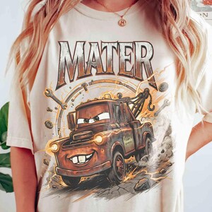 May include: Cream-colored t-shirt featuring a cartoon illustration of Mater from the Cars movie. The design includes the word "MATER" in a distressed font, along with lightning bolts, wrenches, and a speedometer.