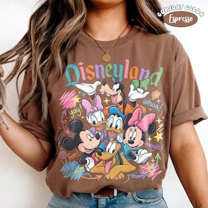 K&ouml;nnte beinhalten: Ein braunes T-Shirt mit einem Grafikdruck von Mickey Mouse, Minnie Mouse, Donald Duck, Daisy Duck, Goofy und Pluto. Der Schriftzug Disneyland ist oben in einer bunten Schrift gedruckt, erg&auml;nzt durch W&ouml;rter wie yay, whoa, magic und fun.