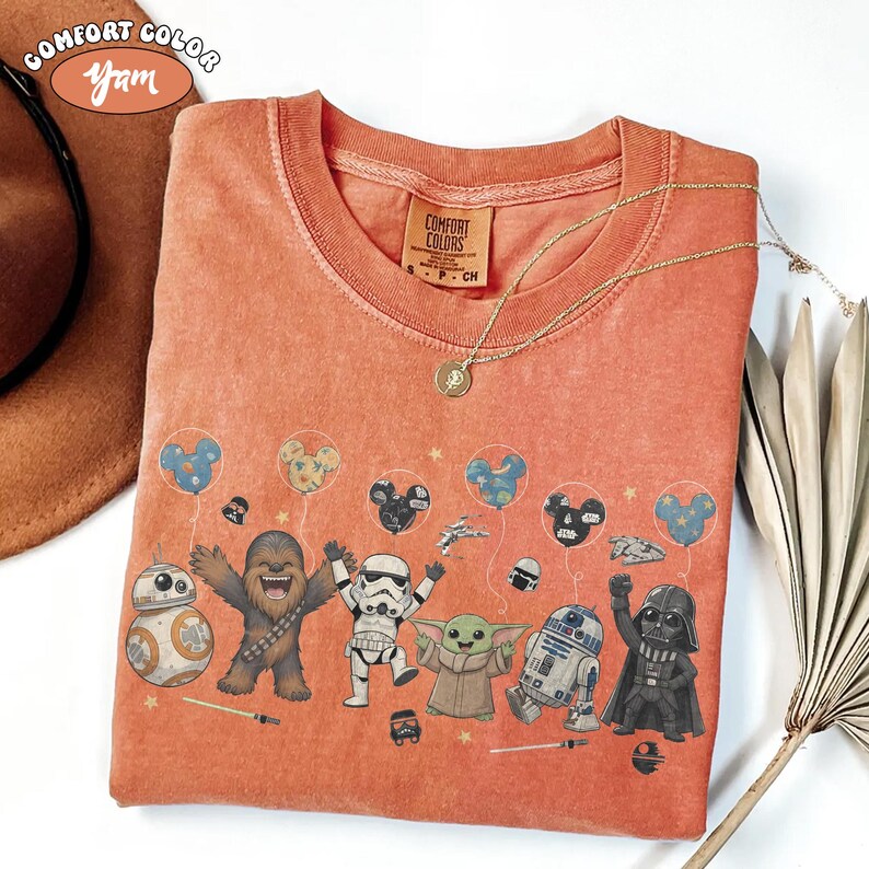 May include: A burnt orange t-shirt featuring a graphic print of Star Wars characters including Chewbacca, a Stormtrooper, Grogu, R2-D2, Darth Vader, and BB-8 holding Mickey Mouse-shaped balloons. The shirt has a crew neckline and a Comfort Colors label.