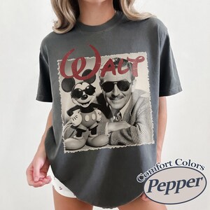 Pu&ograve; includere: T-shirt grigio scuro con grafica vintage di Walt Disney e Mickey Mouse con occhiali da sole. La parola "WALT" &egrave; in corsivo rosso sopra l'immagine. La maglietta ha le parole "Comfort Colors Pepper" stampate.