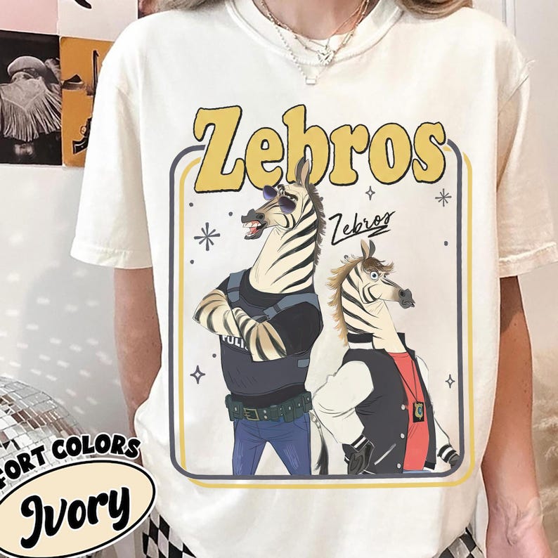 以下が含まれることがあります： 2匹のシマウマの漫画デザインが特徴のアイボリー色のTシャツ。「Zebros」の文字が黄色で描かれ、シマウマはサングラスとジャケットを着用しています。シャツの下部には「Fort Colors」と「Ivory」の文字も表示されています。