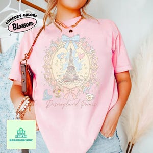Peut inclure: T-shirt rose clair avec un motif Disneyland Paris. Le motif comprend la Tour Eiffel, Mickey Mouse et d'autres &eacute;l&eacute;ments dans un cadre floral. Le texte "Disneyland Paris" est en dessous. Le t-shirt porte &eacute;galement le texte "Comfort Colors Blossom".
