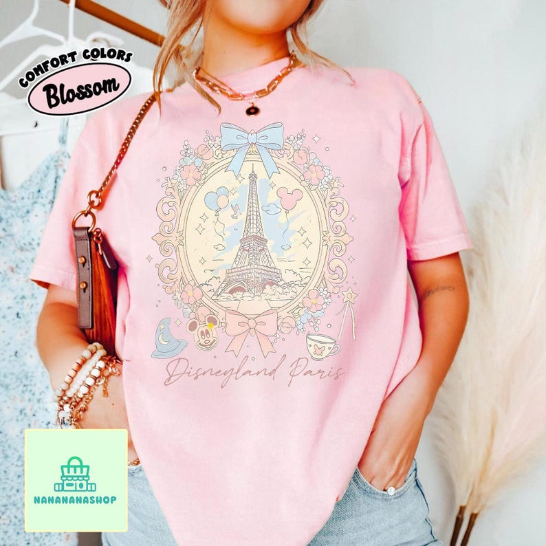 Peut inclure: T-shirt rose clair avec un motif Disneyland Paris. Le motif comprend la Tour Eiffel, Mickey Mouse et d'autres &eacute;l&eacute;ments dans un cadre floral. Le texte "Disneyland Paris" est en dessous. Le t-shirt porte &eacute;galement le texte "Comfort Colors Blossom".