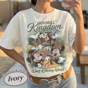 May include: Cream-coloured t-shirt with a Disney character graphic, including Mickey, Minnie, Donald, Daisy, Goofy, and Pluto, with the text "ANIMAL KINGDOM" and "Walt Disney World". The shirt has a crew neck and short sleeves.