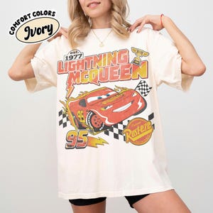 May include: Ivory t-shirt with a Lightning McQueen graphic from the film Cars. The design includes the text "Lightning McQueen" and "95", a checkered flag, and the "Rust-eze" logo. The shirt has a vintage, distressed appearance.