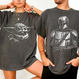 K&ouml;nnte beinhalten: Zwei dunkelgraue T-Shirts. Eines zeigt Grogu mit dem Namen darunter. Das andere zeigt The Mandalorian mit dem Text darunter. Beide Shirts haben kurze &Auml;rmel und eine lockere Passform.
