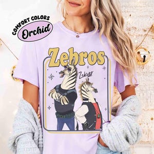 以下が含まれることがあります： 黄色い「Zebros」の文字が入った薄紫色のTシャツ。警察とカジュアルな服装の漫画のシマウマが描かれています。シャツには、「Comfort Colors」と「Orchid」の文字も円形デザインで入っています。