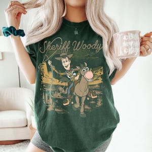 May include: Dark green t-shirt featuring Sheriff Woody from Toy Story. The graphic shows Woody riding a horse with a lasso, set against a desert landscape. The text "Sheriff Woody" is above the image in a script font.