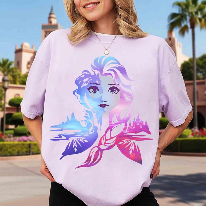 May include: A light purple t-shirt featuring a colorful graphic of a woman's face with blue and purple hair. The design includes a landscape and castle in shades of blue, pink, and purple.
