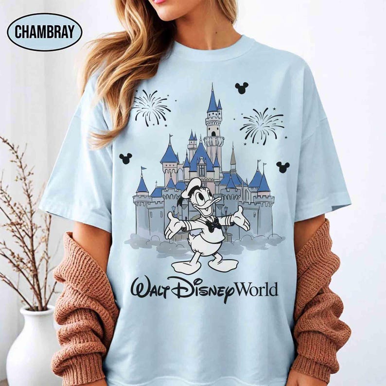 K&ouml;nnte beinhalten: Hellblaues T-Shirt mit einer Grafik von Donald Duck vor einem Schloss und dem Text "Walt Disney World". Das Design beinhaltet Feuerwerk und Mickey Mouse Silhouetten. Das T-Shirt hat eine hellblaue Farbe.