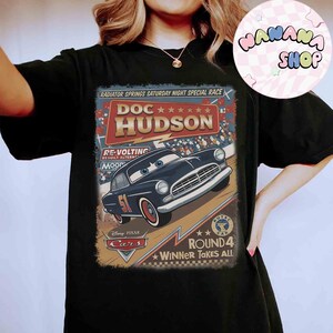May include: Black t-shirt featuring a vintage-style graphic of Doc Hudson from the Disney Pixar Cars movie. The design includes the text "RADIATOR SPRINGS SATURDAY NIGHT SPECIAL RACE" and "ROUND 4 WINNER TAKES ALL".