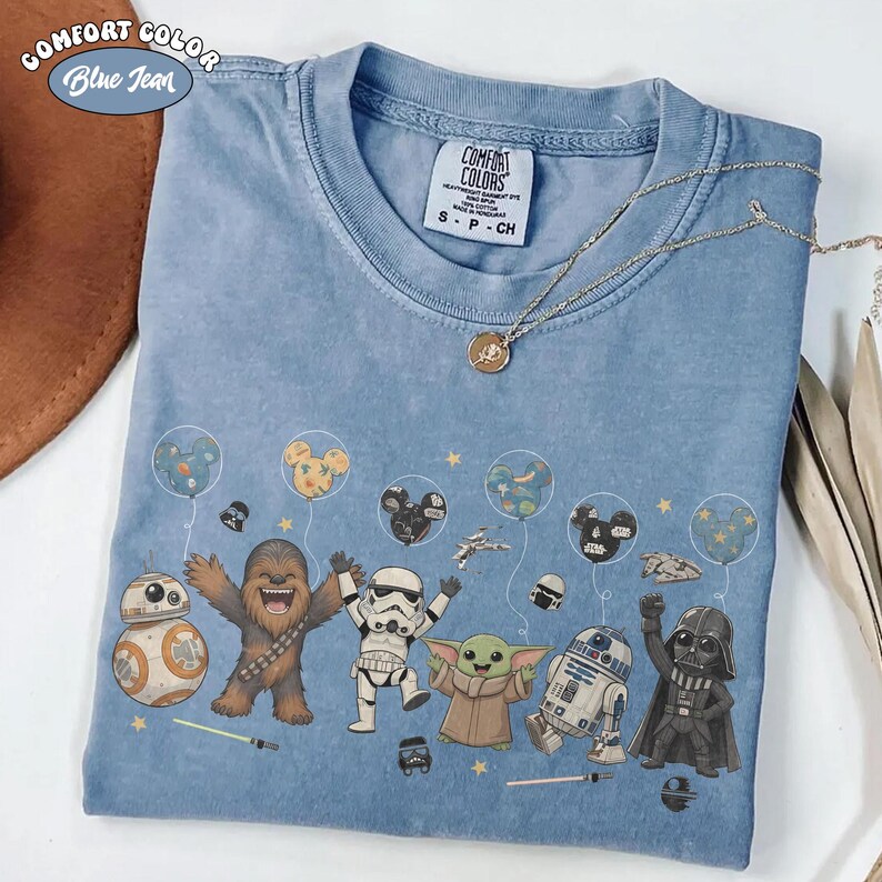 May include: A blue jean colored Comfort Colors t-shirt featuring a graphic print of Star Wars characters including Chewbacca, a Stormtrooper, Grogu, R2-D2, Darth Vader, and BB-8 holding Mickey Mouse shaped balloons and small stars.