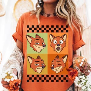 May include: Orange t-shirt with a four-panel cartoon fox design. Each panel shows a different expression against green, orange, and yellow backgrounds. The top and bottom have a black and white checkered border.