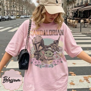Op de afbeelding: Roze T-shirt met een afbeelding van The Mandalorian en Grogu, omringd door bloemen. Het woord "MANDALORIAN" staat bovenaan gedrukt. Een zwarte handtas met kettingriem is zichtbaar.