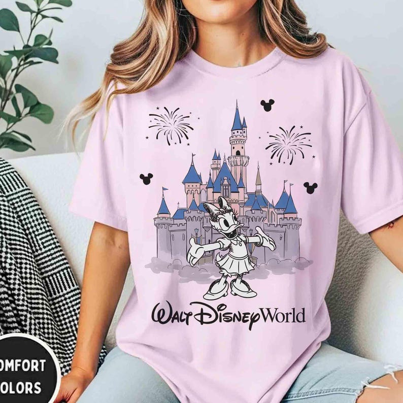 K&ouml;nnte beinhalten: Hellrosa T-Shirt mit einer Grafik von Daisy Duck vor einem Schloss mit blauen T&uuml;rmen und dem Schriftzug "Walt Disney World". Das Design beinhaltet Feuerwerk und Mickey Mouse Silhouetten.