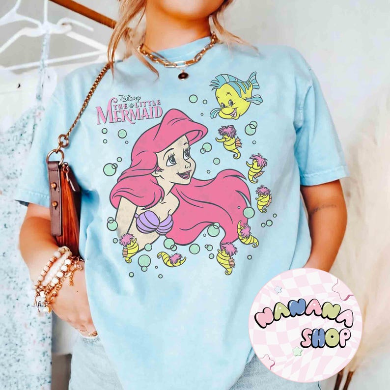 May include: Light blue t-shirt featuring a graphic of Ariel from Disney's The Little Mermaid. Ariel has pink hair and a purple seashell bra. The shirt also includes Flounder and other sea creatures. The text "Disney The Little Mermaid" is printed above Ariel.