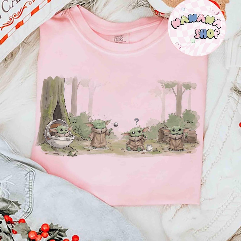 May include: A light pink t-shirt featuring a cartoon illustration of Grogu from The Mandalorian. The design shows Grogu in various poses in a forest setting. The shirt has the text "WANANA SHOP" in a circular logo.