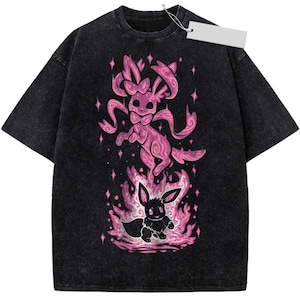 May include: A black acid-wash t-shirt featuring a graphic print of a pink patterned creature floating above a smaller black creature surrounded by pink flames and star accents. A blank white rectangular price tag is attached to the collar.