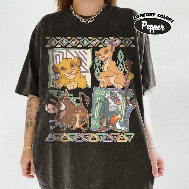 May include: Black t-shirt featuring a colorful graphic of characters from The Lion King. The design includes Simba, Timon, Pumbaa, Rafiki, and Zazu, with geometric patterns. The shirt has the text "Comfort Colors Pepper" in a circular design.