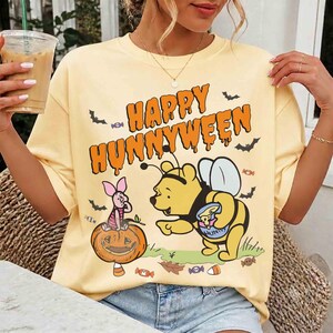 May include: A pale yellow short-sleeve t-shirt featuring a graphic print with the text "HAPPY HUNNYWEEN" in orange. The design includes a yellow bear dressed as a bee, a pink piglet sitting on a jack-o'-lantern, bats, and candy pieces.