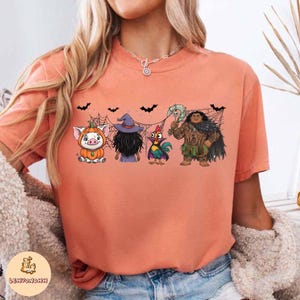 May include: Peach-colored t-shirt featuring a Halloween-themed graphic. The graphic includes cartoon characters dressed in costumes, including a pig in a pumpkin costume, a witch, a rooster, and a character with a weapon.