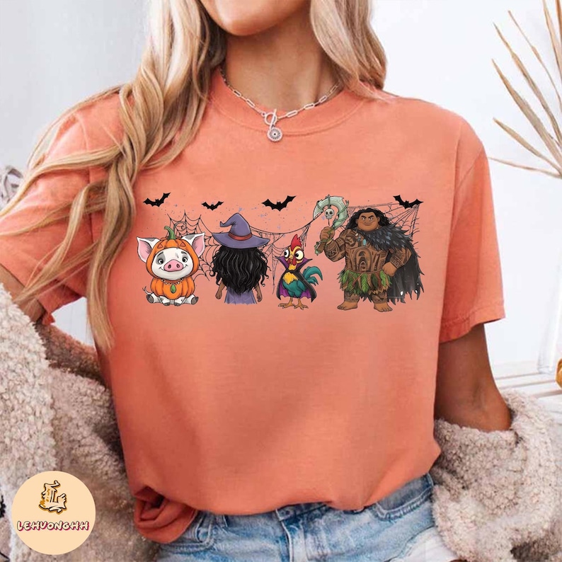 May include: Peach-colored t-shirt featuring a Halloween-themed graphic. The graphic includes cartoon characters dressed in costumes, including a pig in a pumpkin costume, a witch, a rooster, and a character with a weapon.