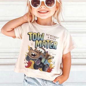 Może przedstawiać: Beżowa koszulka z kr&oacute;tkim rękawem z grafiką postaci Złomka z filmu Pixar Cars. Tekst "TOW MATER" i "OFF WITHOUT A HITCH" jest wydrukowany nad postacią. Koszulka ma okrągły dekolt.