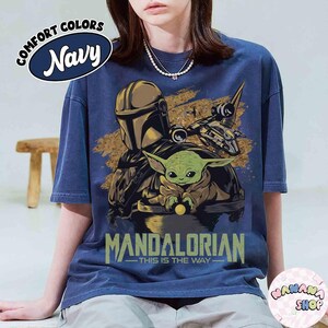 Puede incluir: Camiseta azul marino con un dise&ntilde;o de The Mandalorian y Grogu, con el texto "MANDALORIAN - THIS IS THE WAY". La camiseta tambi&eacute;n tiene el texto "COMFORT COLORS Navy" en un c&iacute;rculo. La camiseta es de corte holgado.
