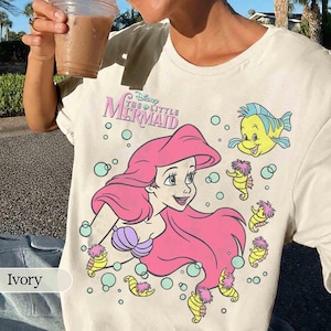 May include: Ivory-colored t-shirt featuring a graphic of Ariel from Disney's The Little Mermaid. The design includes Ariel with pink hair, a purple seashell bra, and Flounder. The shirt also has seahorse and bubble illustrations.