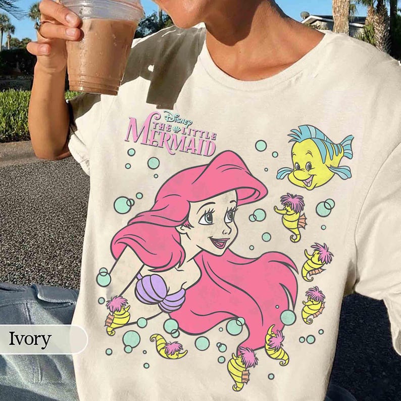May include: Ivory-colored t-shirt featuring a graphic of Ariel from Disney's The Little Mermaid. The design includes Ariel with pink hair, a purple seashell bra, and Flounder. The shirt also has seahorse and bubble illustrations.