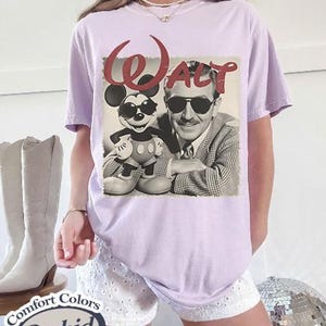 Pu&ograve; includere: Una maglietta lilla Comfort Colors con un'immagine in bianco e nero di Walt Disney e Mickey Mouse con occhiali da sole. La parola "Walt" &egrave; in corsivo rosso sopra l'immagine. La maglietta ha una vestibilit&agrave; rilassata.