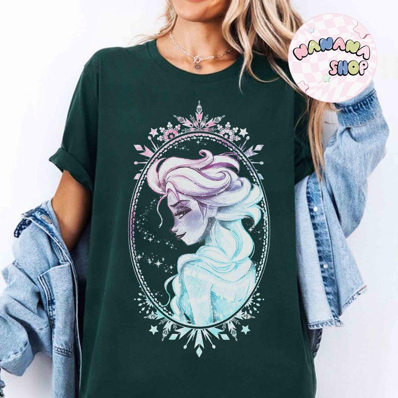 Op de afbeelding: Donkergroen T-shirt met een portret van een vrouw met lichtblauw en paars haar, omlijst in een ovaal met een decoratieve rand. Het shirt heeft opgerolde mouwen en de tekst "NANANA SHOP" in een cirkel.