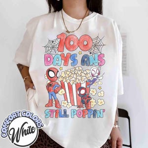 May include: White t-shirt with a colorful graphic featuring Spider-Man characters, popcorn, and the text "100 Days Ans Still Poppin'". The design includes a red, blue, and black color scheme with a playful font. The shirt has a Comfort Colors label.