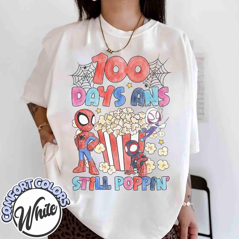May include: White t-shirt with a colorful graphic featuring Spider-Man characters, popcorn, and the text "100 Days Ans Still Poppin'". The design includes a red, blue, and black color scheme with a playful font. The shirt has a Comfort Colors label.