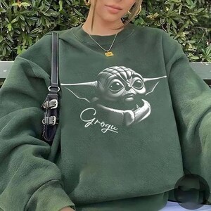 K&ouml;nnte beinhalten: Dunkelgr&uuml;nes Sweatshirt mit einem Grogu-Motiv. Das Design zeigt eine detaillierte Illustration von Grogus Gesicht mit gro&szlig;en Augen und Ohren sowie den Namen "Grogu" in Schreibschrift. Das Sweatshirt hat eine einfarbige gr&uuml;ne Farbe.