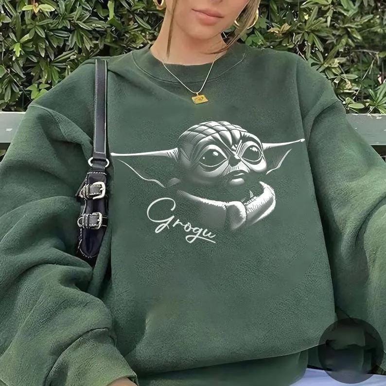 K&ouml;nnte beinhalten: Dunkelgr&uuml;nes Sweatshirt mit einem Grogu-Motiv. Das Design zeigt eine detaillierte Illustration von Grogus Gesicht mit gro&szlig;en Augen und Ohren sowie den Namen "Grogu" in Schreibschrift. Das Sweatshirt hat eine einfarbige gr&uuml;ne Farbe.