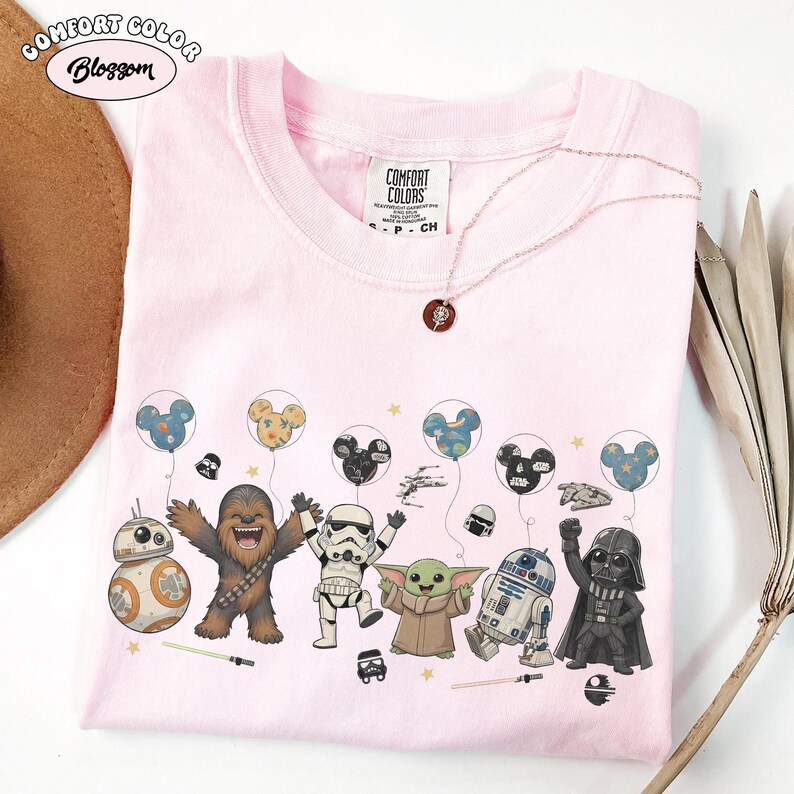 May include: A light pink t-shirt featuring a graphic print of Star Wars characters including Chewbacca, a Stormtrooper, Grogu, R2-D2, and Darth Vader, along with Mickey-shaped balloons, small stars, and lightsabers. The shirt has a Comfort Colors label.
