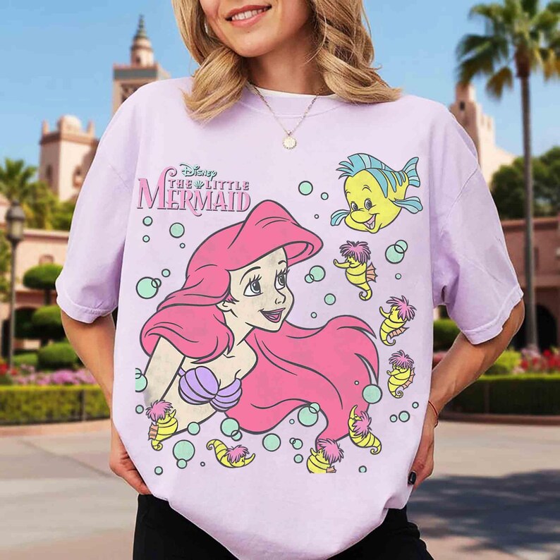 May include: A light purple t-shirt featuring Ariel from Disney's The Little Mermaid. The graphic includes Ariel with pink hair, a purple seashell bra, and Flounder. The shirt also has the text "Disney The Little Mermaid".