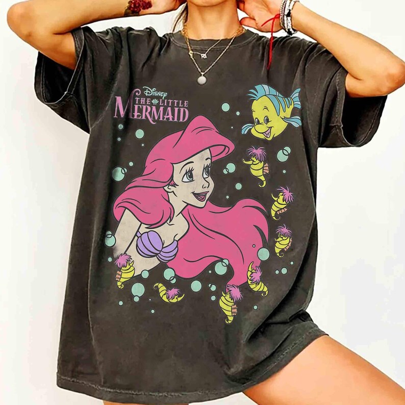 May include: Dark gray t-shirt featuring a graphic of Ariel from Disney's The Little Mermaid. Ariel has pink hair and a purple seashell bra. The shirt also includes Flounder and seahorse illustrations.