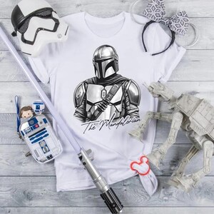 K&ouml;nnte beinhalten: Wei&szlig;es T-Shirt mit einer Schwarz-Wei&szlig;-Illustration von The Mandalorian. Das Bild enth&auml;lt einen Stormtrooper-Helm, ein Lichtschwert, eine R2-D2-Tasche und anderes Zubeh&ouml;r zum Thema Star Wars.